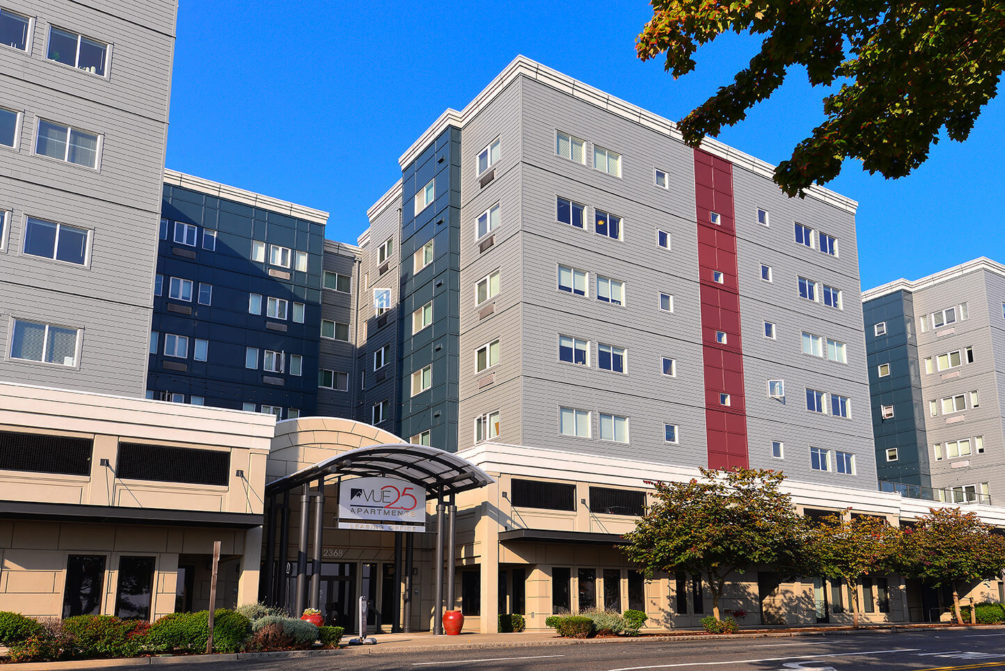 A modern multi-story apartment building with gray siding, red accents, and many windows. An arched entryway with a sign reading “Vue25” is at the front, with trees and a clear blue sky in the background.